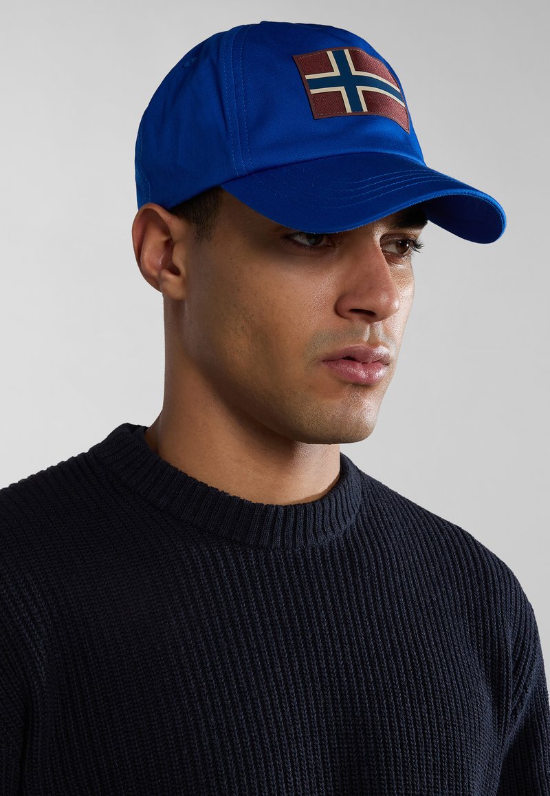 Cappellino blu di tessuto con una toppa rettangolare con design di una bandiera, indossato con un maglione a coste scuro in un primo piano vista di profilo.