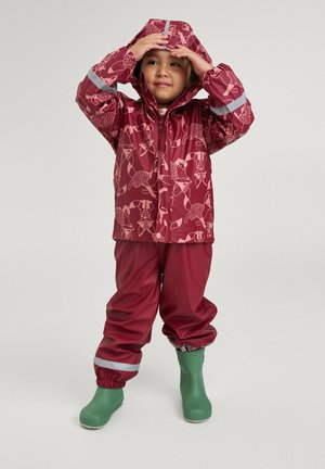 Rote wasserdichte Jacke mit rosa Mustern, elastischen Bündchen und grauen reflektierenden Streifen; kombiniert mit passenden roten Hosen und grünen Gummistiefeln.