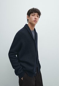 Navy ribbed cardigan with a shawl collar, featuring large buttons and side pockets, paired with dark corduroy pants. Smooth textured fabric.