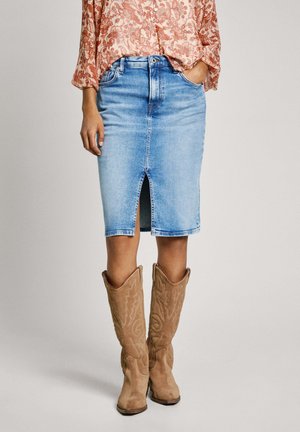 Femme portant une jupe en denim bleu clair longueur genou avec une fente à l'avant, des bottes hautes en daim couleur camel et un chemisier à motif floral.