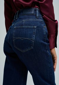 Jeans de mezclilla de color azul oscuro con un corte ajustado. Cuenta con dos bolsillos traseros, costuras visibles y una etiqueta de cuero negro en la cintura.