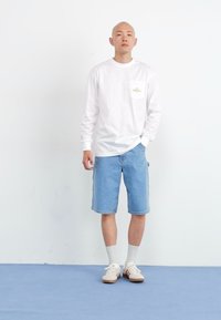 Chemise blanche à manches longues avec une poche poitrine, associée à un short en denim bleu. Des baskets claires et des chaussettes blanches complètent la tenue.