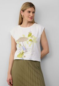 Weißes Baumwoll-T-Shirt mit kurzen Ärmeln, das eine Strandlandschaftsillustration in Grün und Gelb zeigt. Kombiniert mit einem langen, olivgrünen Faltenrock.