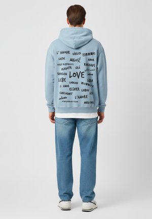 Lichtblauwe hoodie met een capuchon, voorzien van verschillende talen waarin het woord "liefde" in het zwart is gedrukt. Gecombineerd met vervaagde blauwe jeans en witte sneakers.