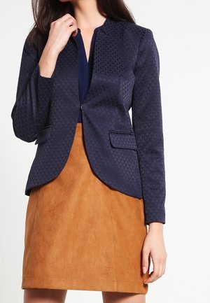 Femme portant un blazer bleu marine texturé sur un haut sombre avec une jupe en daim beige, debout devant un fond blanc uni.
