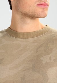 Beige sweatshirt med camo-mønster, med ribbet rund hals, laget av myk stoff; har subtil tekstur og en avslappet passform.