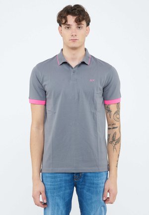 Giovane uomo in piedi, fermo, che indossa una polo grigia con rifiniture rosa e jeans blu, con tatuaggi sul braccio destro.