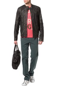 Svart läderjacka med texturerad yta, röd t-shirt med grafiskt tryck, gröna byxor, bär en svart väska och har på sig vita sneakers.