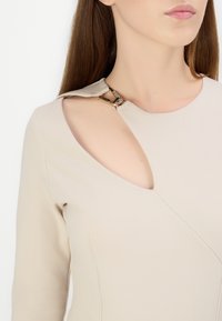 Bež majica dugih rukava s jedinstvenim izrezom na ramenu, s metalnom kopčom ukrašenom rhinestonesom za dodatne detalje. Mekana, teksturirana tkanina.