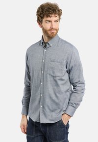 Homme avec des cheveux bouclés et une barbe portant une chemise à boutons grise et un jean foncé, regardant sur le côté, une main dans la poche.