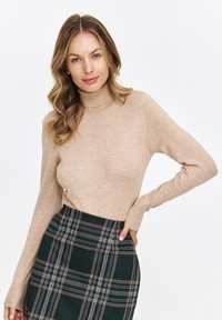 Beżowy sweter z golfem wykonany z miękkiego materiału, z długimi rękawami. Połączony z zieloną spódnicą w kratę z białymi i różowymi akcentami.