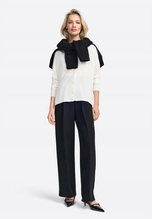 White cardigan with buttons, black textured scarf draped, black wide-leg trousers, and black heeled shoes, standing against a plain background.