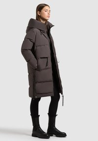 Dlhý páperový kabát tmavosivej farby s vysokým golierom, dvoma prednými vreckami a zipsom. Skombinovaný s čiernymi legínami a hrubými čižmami.