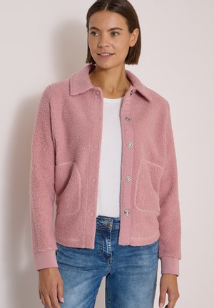 Femme portant une veste texturée rose clair à boutons sur un haut blanc et un jean bleu, debout devant un fond clair uni.