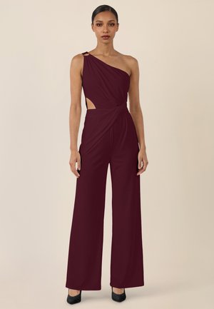 Frau trägt einen ärmellosen, bordeauxroten Ein-Schulter-Jumpsuit mit weitem Bein und seitlichen Ausschnittdetails, stehend vor einem beigen Hintergrund.