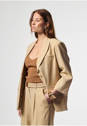 Vrouw met rood haar, in een beige blazer, bijpassende broek en een bruin geribd topje, staand met één hand in de zak tegen een effen achtergrond.