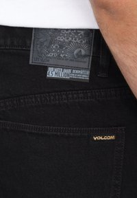 Sorte denim bukser med en tekstureret stof, et dekorativt patch med logo og en label, der angiver vandbesparende egenskaber.