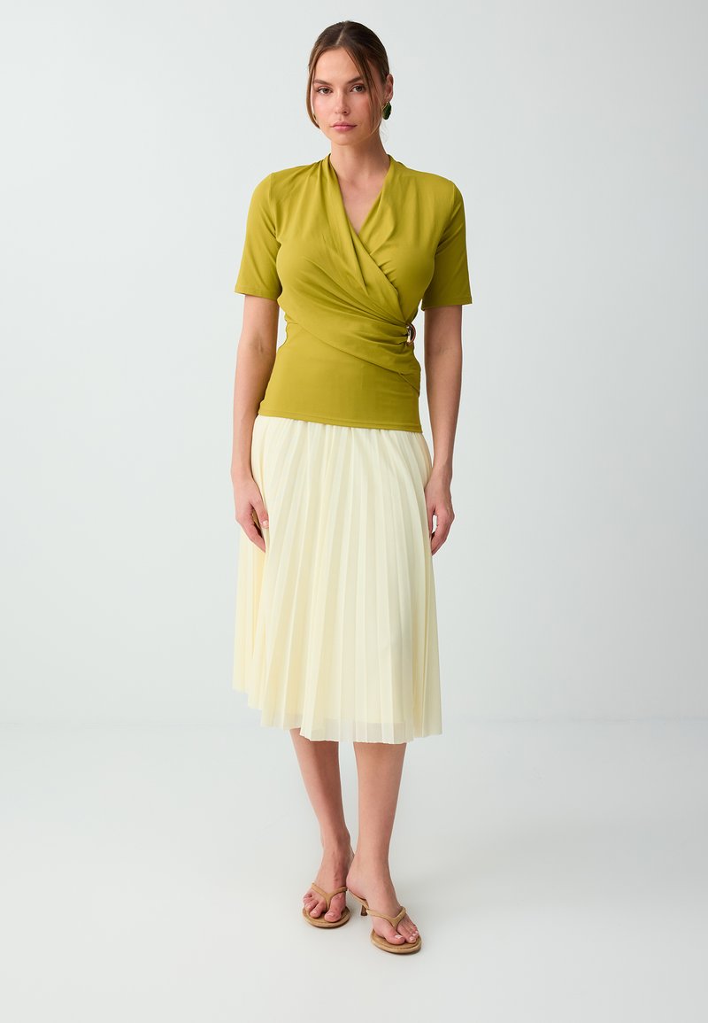 Femme portant un top cache-cœur vert citron à manches courtes et une jupe midi plissée beige, debout devant un fond clair uni.