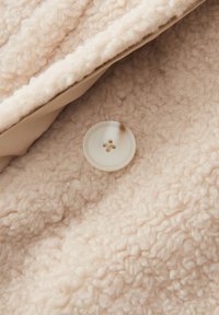 Tessuto beige chiaro con una finitura morbida e testurizzata, caratterizzato da un bottone trasparente con quattro fori. Vista ravvicinata del materiale e dell'hardware.