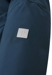 Granatowy materiał o gładkiej teksturze; posiada kwadratową, szaro-rysunkową łatę z wytłoczonym stylizowanym logo "R".
