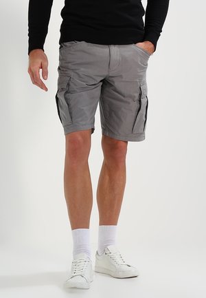 Mann trägt graue Cargoshorts, weiße Socken, weiße Turnschuhe und ein schwarzes Langarmshirt, steht mit einer Hand in der Tasche.