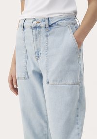 Jasnoniebieskie dżinsy mają luźny krój, duże przednie kieszenie i kontrastowe szwy wzdłuż szwów. Pas wyposażony jest w zapięcie na guzik.