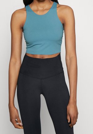 Vrouw draagt een blauwe mouwloze cropped top en zwarte legging met hoge taille, staand tegen een effen lichte achtergrond.