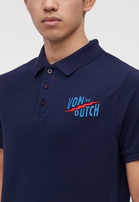 Mann trägt ein marineblaues Polohemd mit drei Knöpfen und dem blau-roten "VON DUTCH ltd" Logo, das auf der Brust aufgestickt ist.