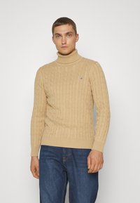 Suéter de punto beige con cuello alto, con puños y dobladillo acanalados, que presenta un pequeño logo en el pecho, combinado con jeans de mezclilla azules.