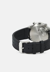 Bracelet de montre en caoutchouc noir avec boucle en métal argenté ; fond de boîtier en acier inoxydable avec détails gravés ; texture lisse et bords arrondis.