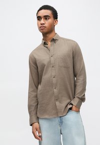 Hnedá košeľa s dlhými rukávmi na zapínanie na gombíky, s jedným náprsníkom a mierne štruktúrovanou tkaninou, v kombinácii so svetlomodrými džínsami.