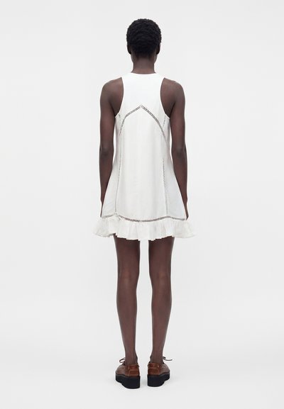 Personne mince debout de dos, portant une robe blanche sans manches avec des détails en dentelle et un ourlet à volants, associée à des chaussures compensées marron.