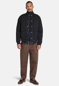 Svart quiltad jacka med tryckknappar, kombinerad med en rutig skjorta och bruna byxor, med en avslappnad passform och fickor. Bruna skor fullbordar looken.