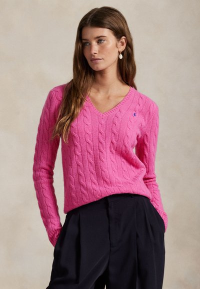 Różowy sweter w splot warkoczowy z dekoltem w serek, o prążkowanej fakturze i małym akcentem w postaci logo. Połączony z czarnymi, wysokim stanem spodniami.