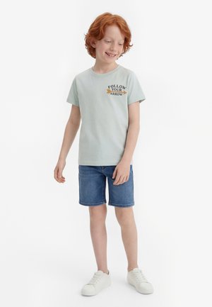 Crvenokosi dječak u svijetloplavoj majici s natpisom "Follow Your Arrow", traper kratkim hlačama i bijelim tenisicama, stoji i gleda dolje.