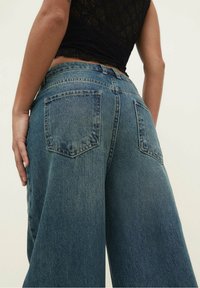 Jeans de mezclilla de tiro alto y pierna ancha en azul medio con un acabado desgastado. Cuenta con dos bolsillos traseros y costuras visibles.