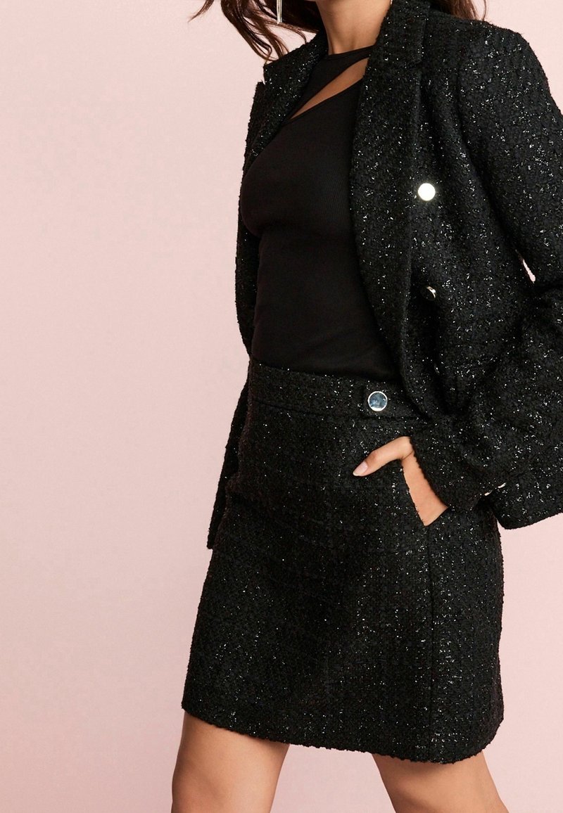 Schwarzer strukturierter Blazer mit metallischen Knöpfen, kombiniert mit einem taillierten schwarzen Rock. Das Outfit zeichnet sich durch einen dezenten Glanz und einen modernen Schnitt aus.