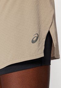 Beežidest sportlikud lühikesed püksid tekstuurse mustriga, külgsise lõikega ja halli Asicsi logoga alumisel kohal. Must sisekatte osa on nähtav.