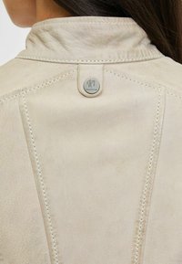 Giacca in pelle beige chiaro con colletto alla mandarino, cuciture a contrasto e una targhetta metallica con logo sulla parte posteriore. Texture liscia con una leggera lucentezza.