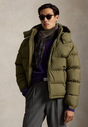 Homme portant des lunettes de soleil foncées, une doudoune verte, un pull violet, un pantalon gris et une écharpe à carreaux sur un fond uni.