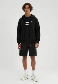 Sudadera negra con cremallera y capucha, combinada con una camiseta negra con rayas blancas, pantalones cortos negros y zapatillas blancas. Tela suave, diseño casual.