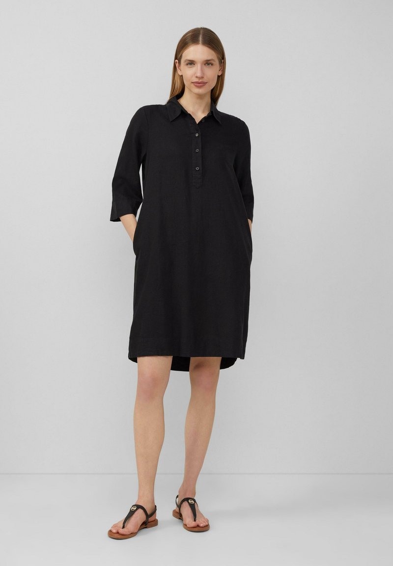 Schwarzes Leinenkleid mit Kragen, dreiviertel Ärmeln, Knopfverschluss und seitlichen Taschen; das Model trägt braune Sandalen.