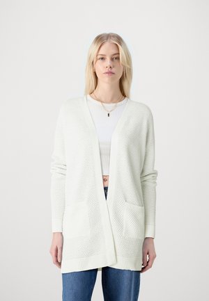 Lichte, textuurrijke, open voorkardigan in crème met een gebreid patroon, lange mouwen en twee voorzakken. Draagt over een witte top.