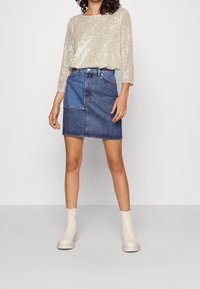 Haut beige à paillettes avec manches trois-quarts, associé à une jupe en jean bleue avec un design patché. Des bottines blanches complètent le look.