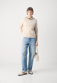 Pull en laine beige avec un col texturé, jeans bleu clair et chaussures beiges avec des accents noirs. Tient un petit sac à main beige clair.