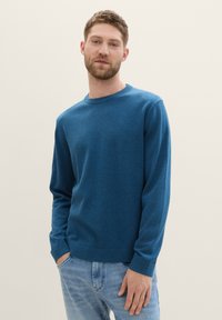 Pulover albastru cu guler rotund, dintr-un material moale, cu mâneci lungi și mansete cu riburi. Purtați cu blugi bleu deschis.