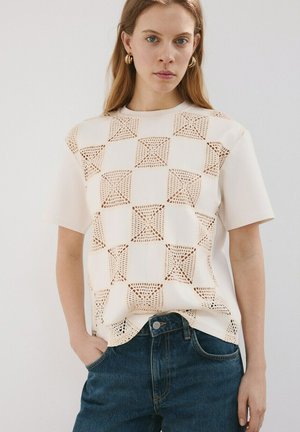 Jeune femme portant un top crème à manches courtes avec des motifs carrés au crochet et un jean bleu, debout, une main dans la poche.