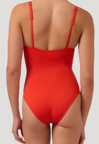 Kvinna som bär en klar röd baddräkt med tunna justerbara axelband, visad från sidan mot en neutral ljus bakgrund.