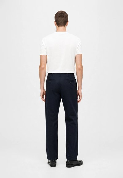 Homme debout, de dos, portant une chemise blanche unie à manches courtes, un pantalon bleu marine et des chaussures noires devant un fond clair uni.