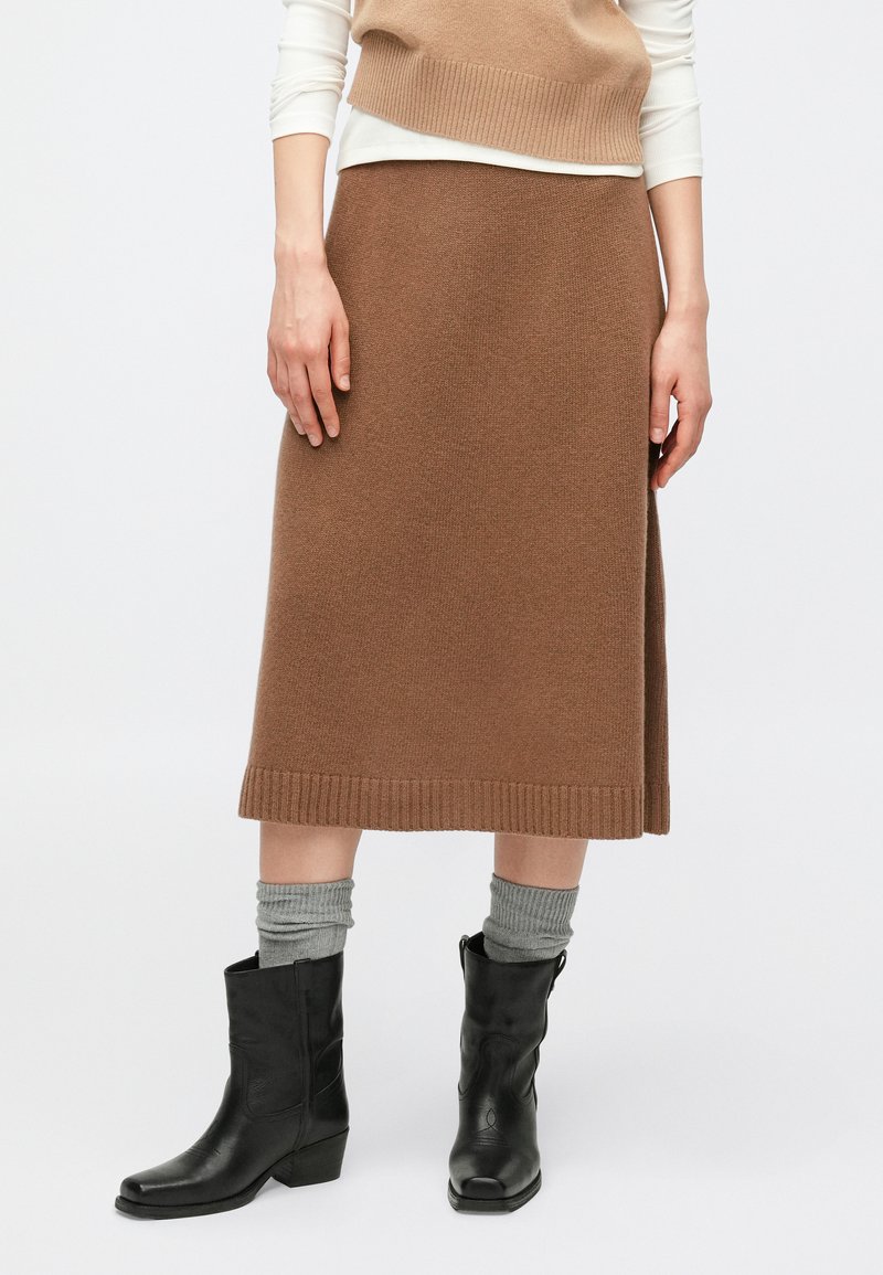 Καφέ πλεκτή μίντι φούστα με ριμπ τελείωμα, συνδυασμένη με γκρι κάλτσες και μαύρα μποτάκια αστραγάλου. Το σύνολο περιλαμβάνει ένα στρωμένο τοπ σε ουδέτερους τόνους.